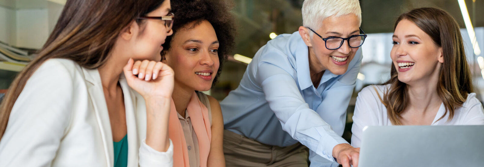 Eine ältere Geschäftsfrau zeigt drei jüngeren Geschäftsfrauen etwas an einem Laptop. Die Stimmung ist freudig.