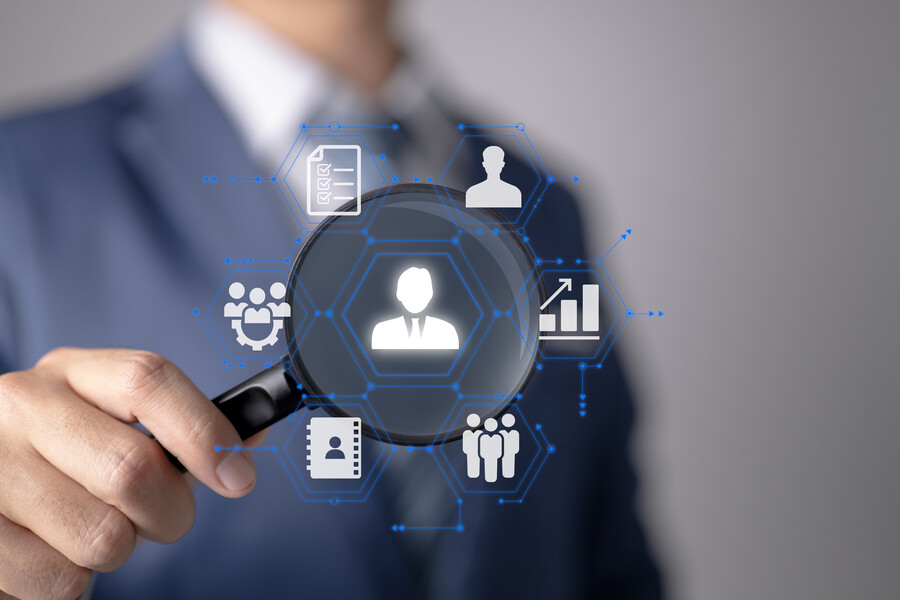 Nahaufnahme einer männlichen Hand, die eine Lupe hält. Im Fokus dieser sind verschiedene Business-Icons z. B. Diagramme, Personen und Dokumente.