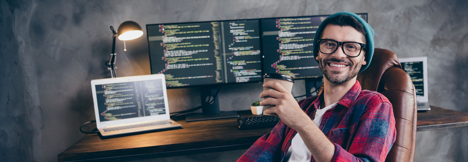 Lässig gekleideter, junger Programmierer mit Brille, Bart und Mütze sitzt zurückgelehnt in seinem Bürostuhl vor seinem Schreibtisch, wobei er einen Kaffeebecher in der Hand hält und zufrieden in die Kamera lacht.