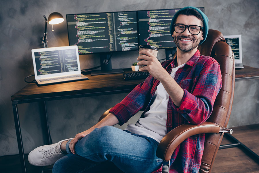 Lässig gekleideter, junger Programmierer mit Brille, Bart und Mütze sitzt zurückgelehnt in seinem Bürostuhl vor seinem Schreibtisch, wobei er einen Kaffeebecher in der Hand hält und zufrieden in die Kamera lacht.