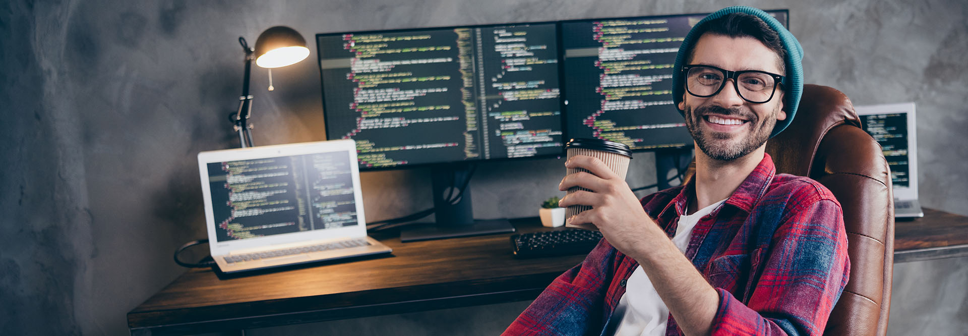 Lässig gekleideter, junger Programmierer mit Brille, Bart und Mütze sitzt zurückgelehnt in seinem Bürostuhl vor seinem Schreibtisch, wobei er einen Kaffeebecher in der Hand hält und zufrieden in die Kamera lacht.