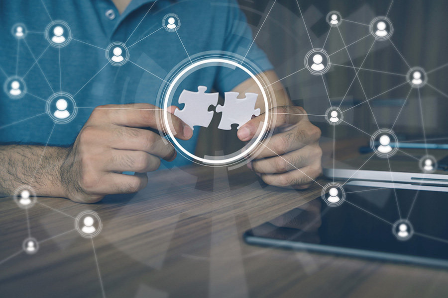 Ein Mann steckt zwei passende Puzzle-Teile ineinander. Auf einer virtuellen Ebene davor spannt sich ein Netzt aus verbundenen Personen-Icons. Darin ist ein Kreis integriert, der den Fokus auf die Puzzle-Teile legt.