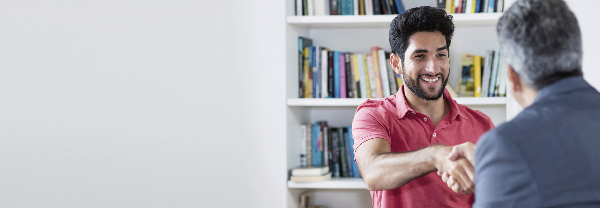 Junger Mann mit Migrationshintergrund sitzt einem älteren Herrn in einem Büro gegenüber, der ihm die Hand gibt