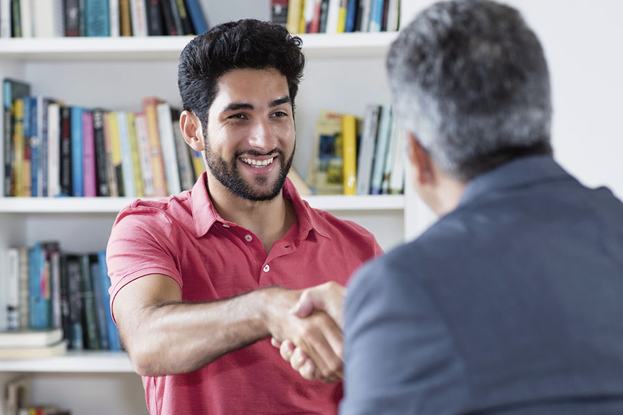 Junger Mann mit Migrationshintergrund sitzt einem älteren Herrn in einem Büro gegenüber, der ihm die Hand gibt