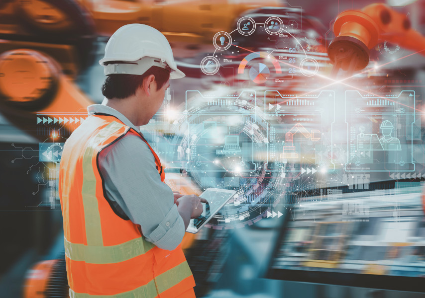 Industriearbeiter in mit Warnweste und Helm von hinten, der über ein Tablet Roboterarme steuert.