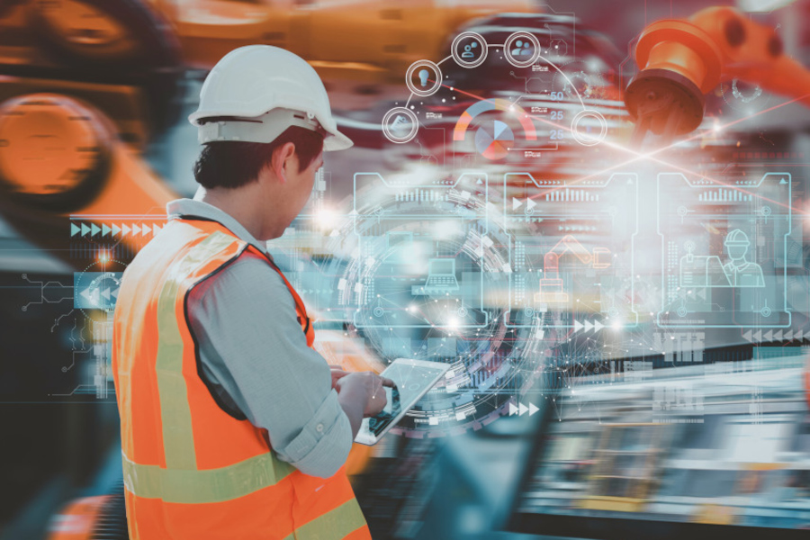 Industriearbeiter in mit Warnweste und Helm von hinten, der über ein Tablet Roboterarme steuert.