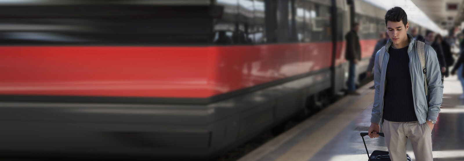 A young, dark-haired man is walking along a train parked on the platform, pulling a rolling suitcase. He is looking down.