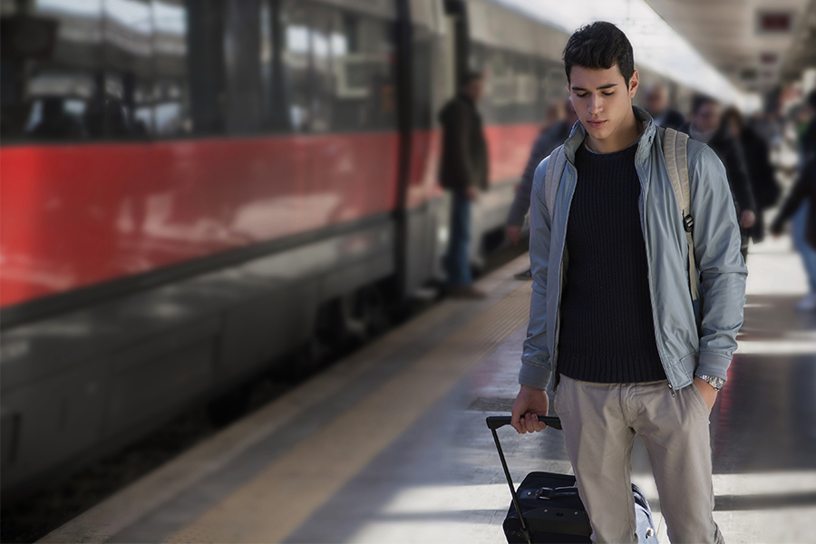A young, dark-haired man is walking along a train parked on the platform, pulling a rolling suitcase. He is looking down.