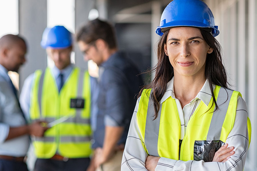 Portrait of a female architect standing on a construction site, smiling confidently. In the background, a group of men are looking at a plan. The focus is on the woman.
