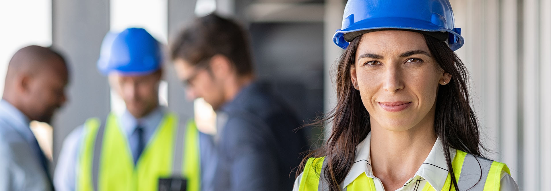 Portrait of a female architect standing on a construction site, smiling confidently. In the background, a group of men are looking at a plan. The focus is on the woman.