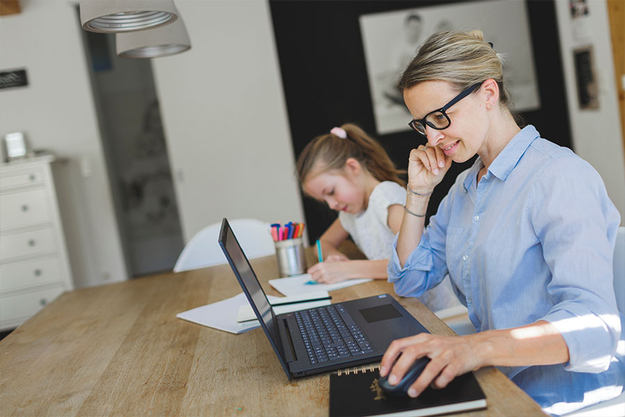 Junge Mutter sitzt zu Hause am Tisch und arbeitet an einem Laptop. Dahinter am selben Tisch sitzt Ihre kleine Tochter und malt.