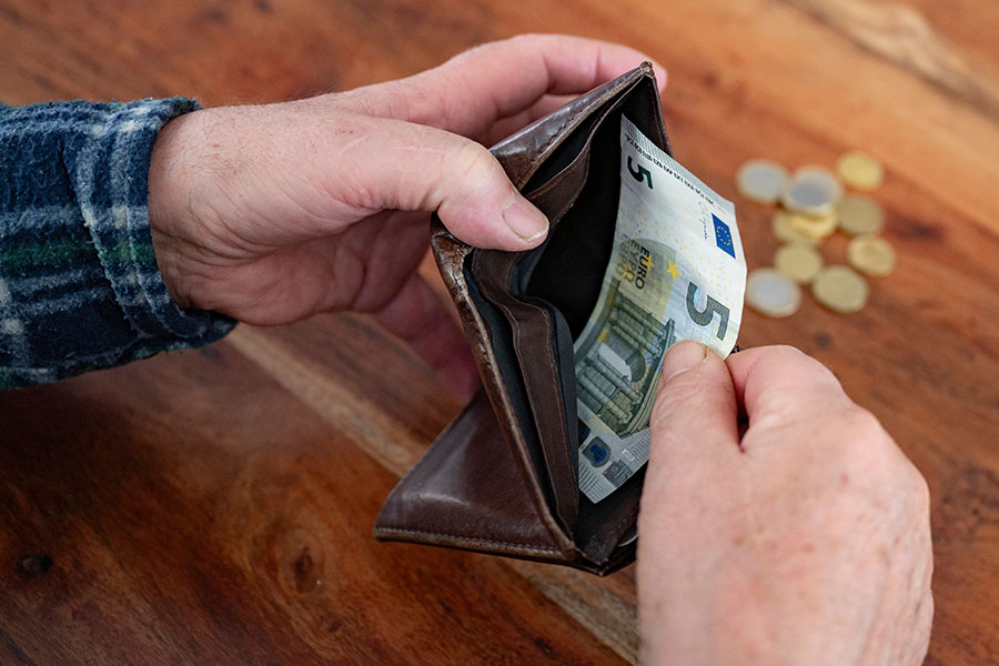 Ein Mann hält seinen Geldbeutel in der einen Hand und zieht mit der anderen den letzten 5-Euro-Schein heraus. Im Hintergrund liegen einige Münzen auf dem Tisch.