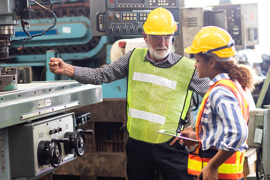Ein älterer grauhaariger Arbeiter erklärt einer jungen Arbeiterin die Funktion einer Metalldrehmaschine.