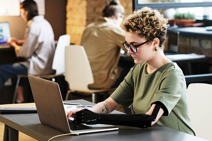Junge Frau mit Brille und Armprothese arbeitet im Büro an einem Laptop.